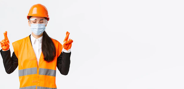 Covid19 safety protocol enterpise construction preventing virus concept hopeful asian female engineer helmet face mask praying close eyes cross fingers good luck white background 1258 95995